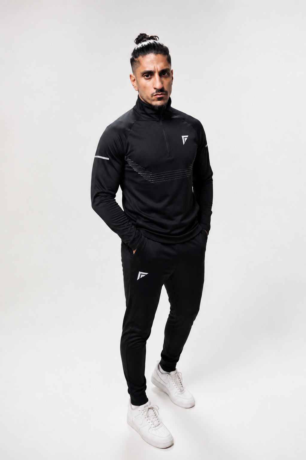 A man modeling a black athletic half-zip pullover and matching black joggers with white sneakers, standing against a light gray studio background.